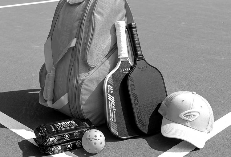 A set of pickleball gear including three paddles, two yellow perforated balls, an orange and blue duffel bag, and a black cap, all branded with the Gamma logo.