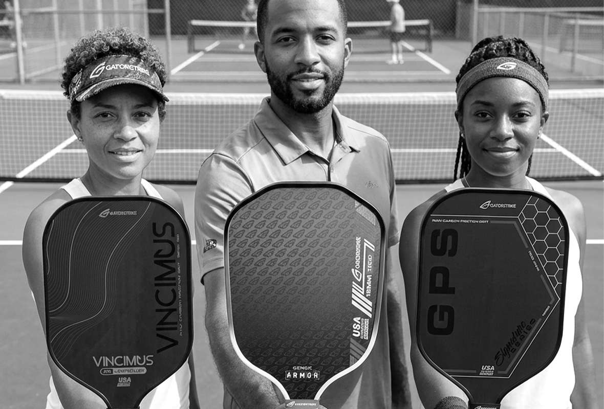 Three pickleball paddles stand upright side by side. Each paddle has a unique design: one is red and gray, one is black with gold text, and one is black with blue accents. All have white handles.