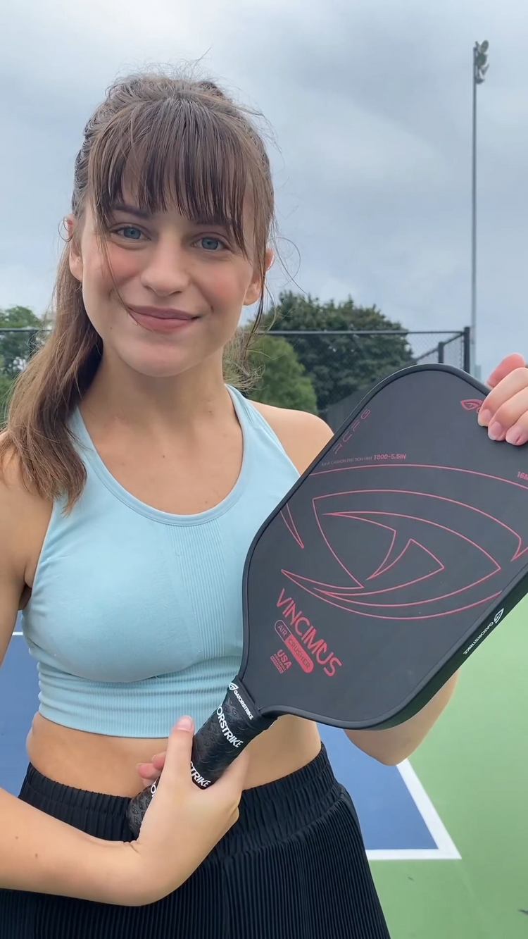 Young woman smiling on the court, wearing a light blue sports bra, holding a pickleball paddle and official pickleball rules.