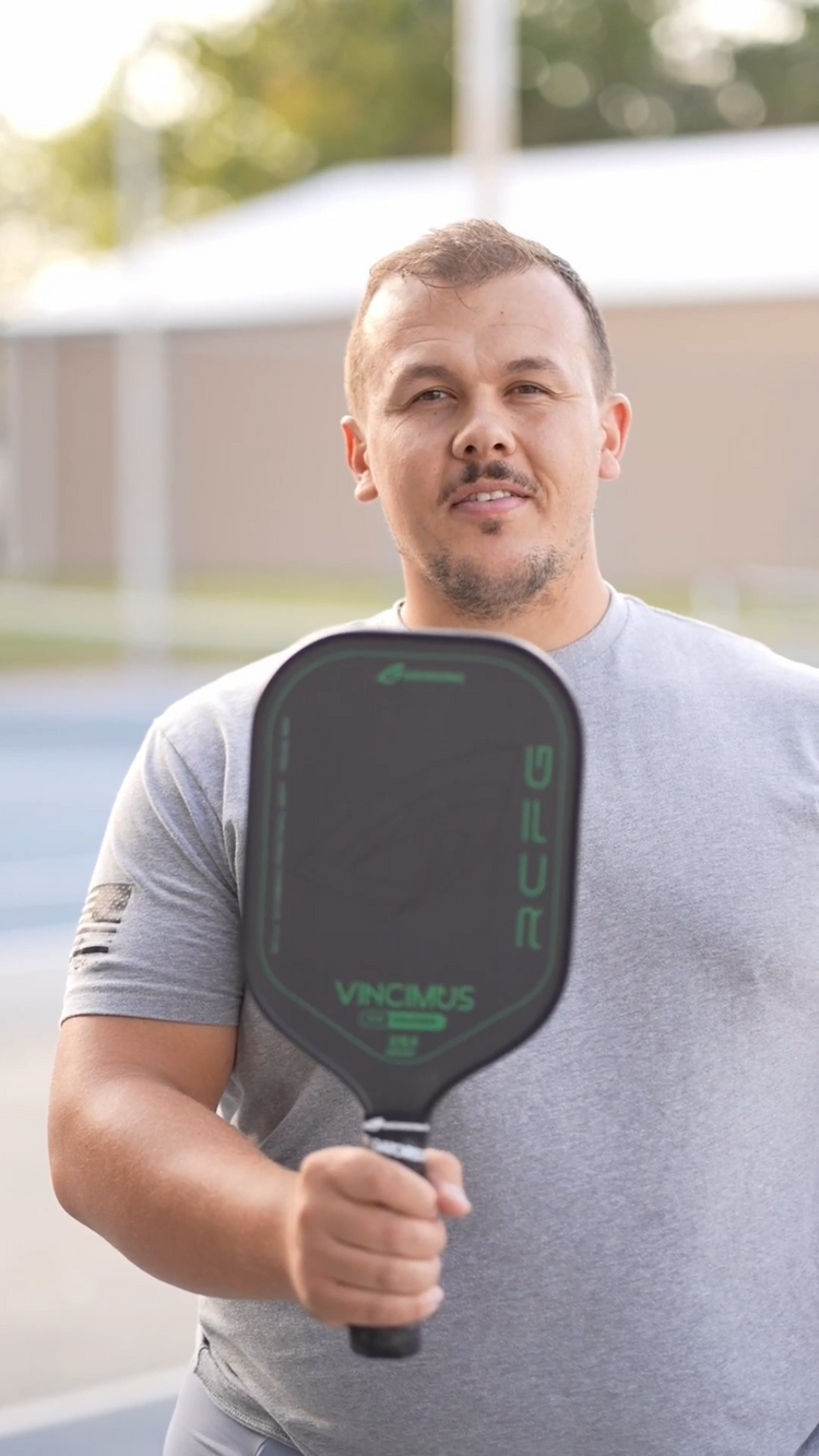Man in a gray shirt holding a black pickleball paddle and a pickleball rulebook, standing on an outdoor court, looking at the camera.