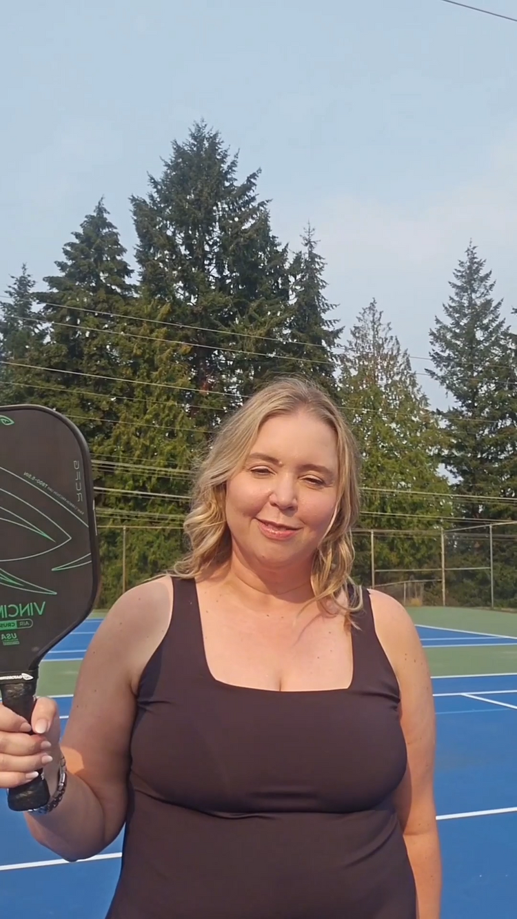 Woman holding a pickleball paddle on an outdoor court, GatorStrike pickleball manual in hand, with trees and clear sky.