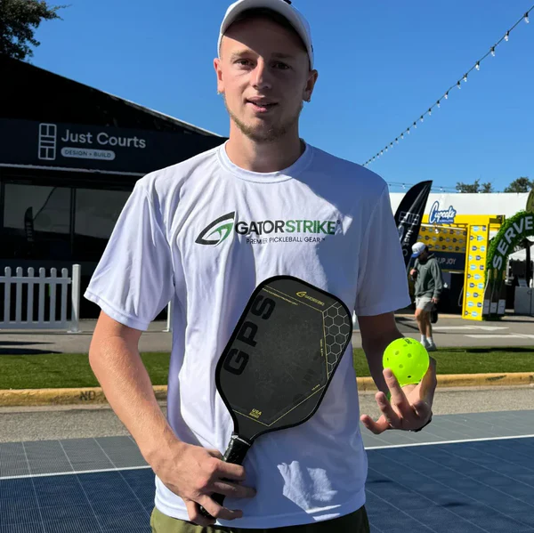 A young man in a white Gator Strike shirt and white cap stands on an outdoor pickleball court, holding a black pickleball paddle and a yellow pickleball. Behind him are court fences, signs, and people walking by on a sunny day.