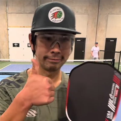 Man wearing cap and goggles gives a thumbs up, holding a pickleball paddle and the official pickleball rules on an indoor court.