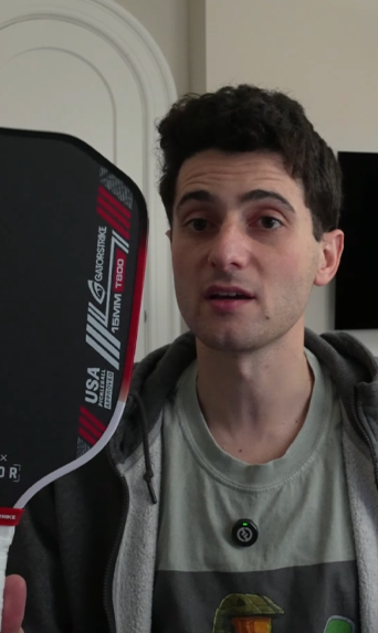 A man indoors holds a black pickleball paddle with red and white stripes, wearing a graphic gray shirt; see official pickleball rules.