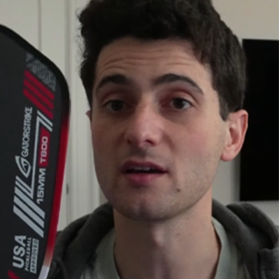 A man with dark hair holds a black and red pickleball paddle, referencing the pickleball rulebook indoors.