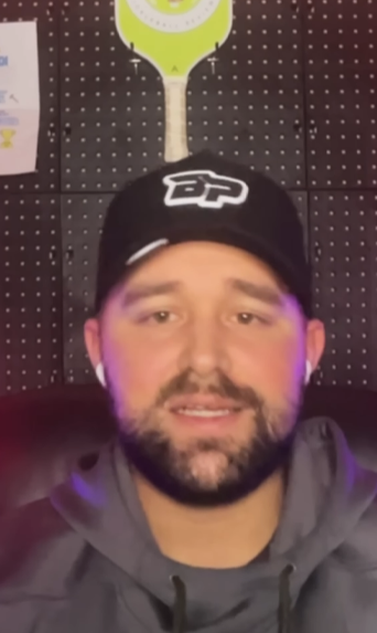 A bearded man in a black cap and gray hoodie sits indoors, AirPods in, with a GatorStrike pickleball manual on the wall behind him.