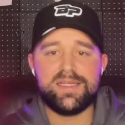 A bearded man in a black hat with a white logo wears AirPods and a gray hoodie, reading the official pickleball rules indoors.