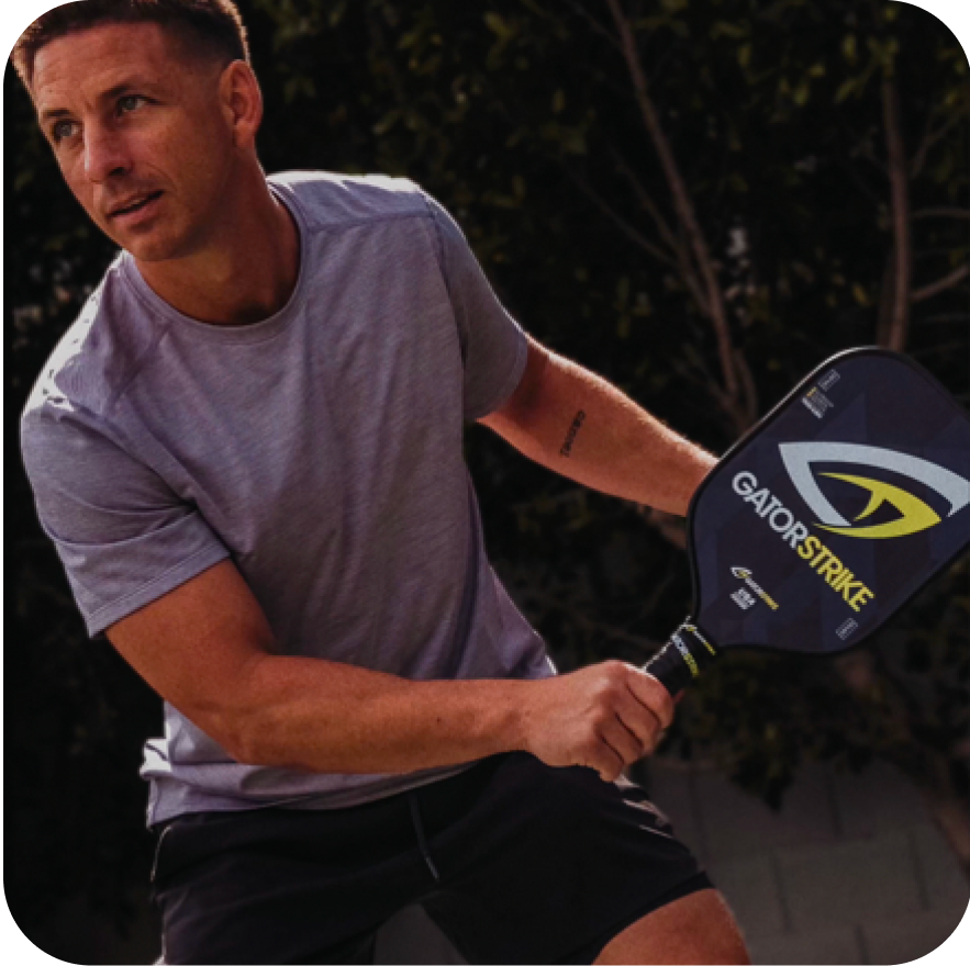 A man in a light purple athletic shirt and black shorts holds a GatorStrike pickleball paddle, preparing to hit a ball. He is outdoors, with trees and a fence in the background, looking focused and ready to play.