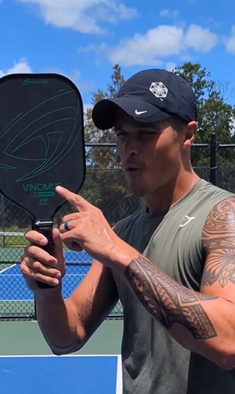Man in a cap points at a pickleball paddle, showing a tattooed arm—ready to consult the pickleball rulebook on an outdoor court.