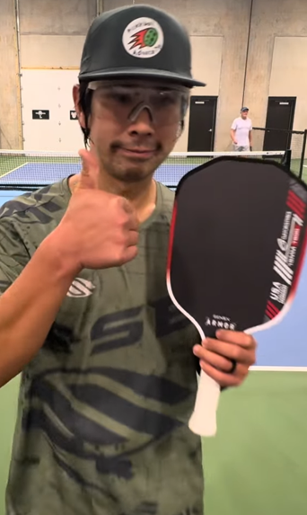 Person holding a pickleball paddle and giving a thumbs-up on a pickleball court.