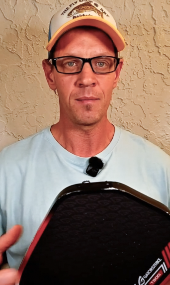 Man in glasses, light blue shirt, and yellow cap holds a black pickleball paddle with a GatorStrike pickleball manual nearby.