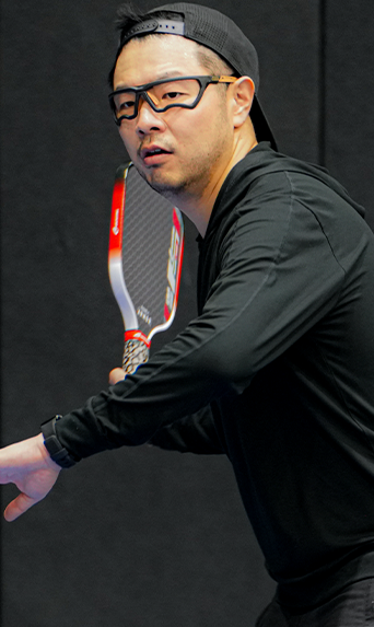 Man in black sportswear and protective glasses holds a racquet, ready to hit a ball indoors, following the pickleball rulebook.