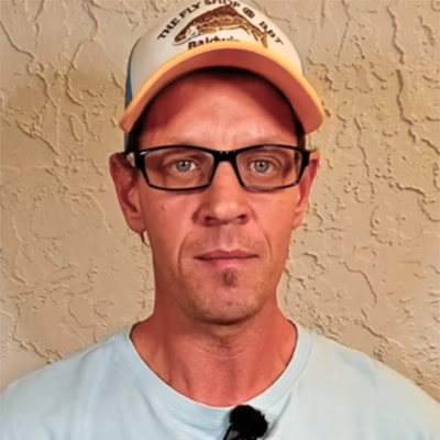 Man with glasses and a cap in a light blue shirt stands by a beige wall, holding a pickleball rulebook.