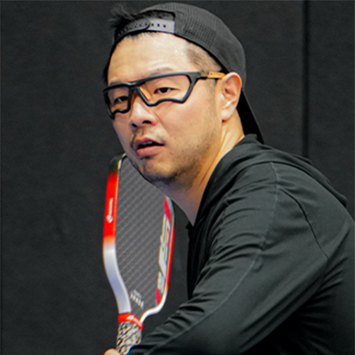 A man in a backward cap and goggles holds a racquet, ready to play by the official pickleball rules against a dark background.